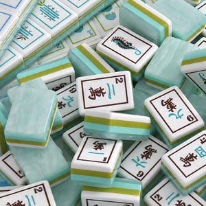 Pode incluir: Uma cole&ccedil;&atilde;o de pe&ccedil;as retangulares de mahjong com face branca, caracteres chineses em preto e azul-petr&oacute;leo, n&uacute;meros e ilustra&ccedil;&otilde;es de um cavalo-marinho e bambu. As pe&ccedil;as t&ecirc;m um verso em azul claro marmorizado e bordas verdes.