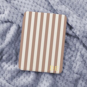 Könnte beinhalten: Ein rechteckiges Etui mit vertikalen Streifen in Beige und Hellbraun. Goldfarbene Initialen "DE" sind in der unteren rechten Ecke zu sehen. Das Etui liegt auf einer weichen, hellblauen, strukturierten Oberfläche.