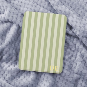 Könnte beinhalten: Eine rechteckige Hülle mit vertikalem Streifenmuster in Hellgrün und Off-White. Die goldenen Initialen "DE" befinden sich in der unteren rechten Ecke. Die Hülle liegt auf einer weichen, strukturierten, hellgrauen Oberfläche.