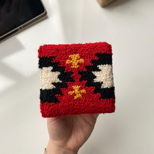 May include: A red, black, white, and yellow patterned square textile. The textile has a textured, looped surface. The design features geometric shapes and floral-like accents. The item is held in a hand.
