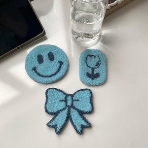 Pode incluir: Três bases para copos azuis texturizadas com bordas azuis escuras. Uma é redonda com um rosto sorridente, outra é retangular com uma flor e a terceira tem formato de laço. Um copo de água está por perto.