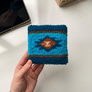 Puede incluir: Un posavasos cuadrado y texturizado con un diseño geométrico turquesa y marrón. El posavasos presenta una forma de diamante central y rayas horizontales, todo sobre un fondo azul verdoso oscuro. Un posavasos es sostenido por una mano.