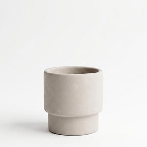 Puede incluir: Una maceta cilíndrica de color beige claro con un acabado mate suave. El recipiente presenta un diseño escalonado distintivo con una sección superior más ancha que se estrecha hacia una base más angosta, creando una silueta estructurada.