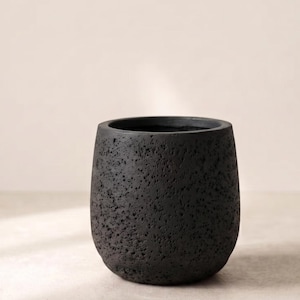 Puede incluir: Una maceta cilíndrica con un acabado en negro carbón oscuro y una superficie muy texturizada y porosa. El recipiente presenta una parte superior ancha y abierta y una base redondeada, mostrando un aspecto rugoso similar a la piedra.