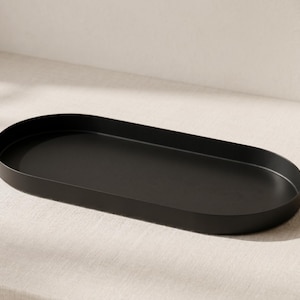 May include: A matte black oval tray with a smooth, continuous rim and a flat base. The tray features a simple, elongated shape with rounded ends, providing a clean and functional surface for organizing small items or displaying decorative objects.