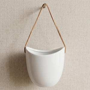 May include: A white ceramic wall planter with a smooth, glossy finish and a curved, semi-circular opening. The planter is suspended by a thin, light brown leather strap that forms a triangular loop, allowing it to be hung from a wall hook.