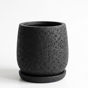 May include: A black ceramic plant pot with a matching saucer. The pot has a textured, porous surface and a rounded, slightly tapered shape. The dark colour and unique texture give it a modern, minimalist aesthetic. Suitable for indoor or outdoor use.