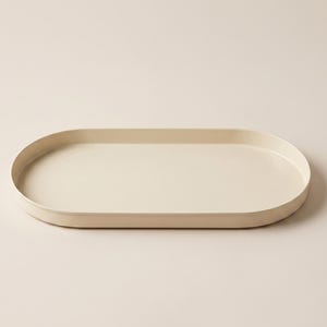 Può includere: Un vassoio di forma ovale in un colore crema solido. Il vassoio presenta un bordo liscio e continuo e una base piatta, offrendo un aspetto pulito e uniforme. Il design è semplice e funzionale, con una finitura opaca su tutta la superficie.