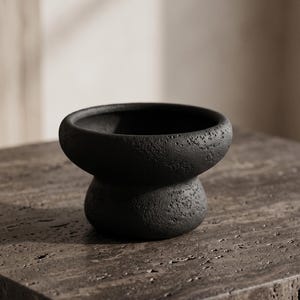 May include: A small, matte black bowl with a textured, porous surface. The bowl features a wide, rounded upper rim that tapers down into a narrower, bulbous base, creating a distinct hourglass-like silhouette with a deep, open interior cavity.