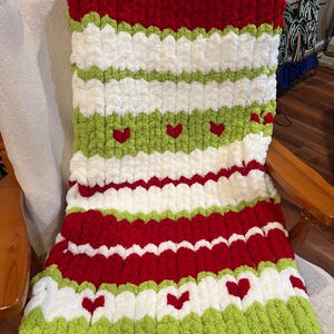 May include: A chunky knit blanket featuring horizontal stripes in red, white, and lime green. The blanket is constructed with thick, soft yarn and includes small red heart motifs integrated into the white and green striped sections.