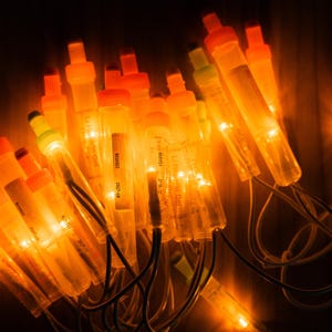 May include: A string of illuminated orange tube lights featuring translucent cylindrical casings with orange caps. Each light unit contains a small glowing bulb, connected by thin dark wires, creating a warm, bright, and clustered display.