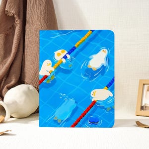 Könnte beinhalten: Ein rechteckiges Notizbuch mit einer Illustration eines leuchtend blauen Schwimmbeckens auf dem Einband. Das Motiv zeigt mehrere weiße Cartoon-Enten, die im Wasser schwimmen und treiben, getrennt durch rote, gelbe und blaue Bahnenmarkierungen.