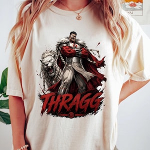 May include: Cream-colored t-shirt featuring a graphic of a superhero with a lion, and the word "THRAGG" in red. The superhero is depicted in a red and white outfit with a red cape. The lion is white with a fierce expression.