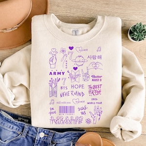 Może przedstawiać: Kremowa bluza z fioletowymi wzorami graficznymi i tekstem. Projekt zawiera serca, kwiaty, gesty dłoni oraz słowa "ARMY", "BTS", "HOPE", "NEVERMIND", "Butter", "The Best of K-Pop" i "2026 World Tour."