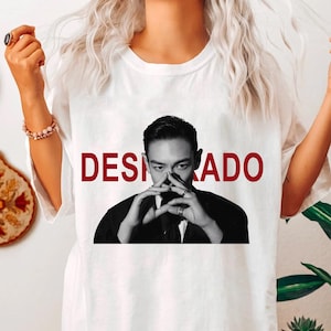 May include: White t-shirt with the word "DESPERADO" in red block letters. The shirt features a black and white image of a man with his hands near his face. The shirt is a casual style.