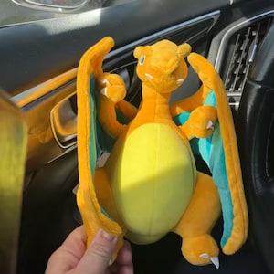 May include: A plush Charizard toy in shades of orange, yellow, and teal. The stuffed animal has large wings, a long tail, and a friendly expression. The toy is held in a car.