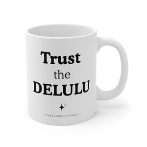 Peut inclure: Un mug en céramique blanc avec le texte imprimé en noir Trust the DELULU dans une police serif grasse. Sous le texte se trouve une petite icône d'étoile à quatre branches avec des points et le nom PIXELSENSHI STUDIO en police sans-serif.
