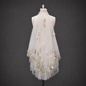 May include: A sheer, off-white tulle veil featuring delicate, multi-colored floral embroidery and small, scattered fabric flowers. The veil is gathered at the top with a small floral accent and drapes downward in a layered, semi-transparent design.
