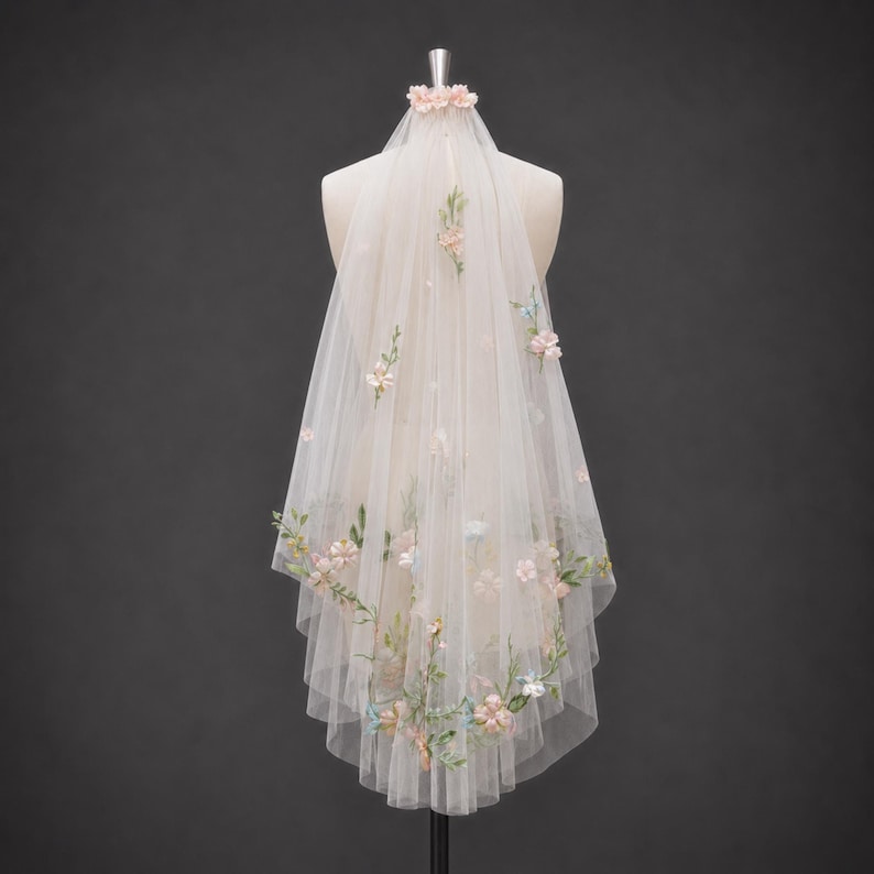 May include: A sheer, off-white tulle veil featuring delicate, multi-colored floral embroidery and small, scattered fabric flowers. The veil is gathered at the top with a small floral accent and drapes downward in a layered, semi-transparent design.