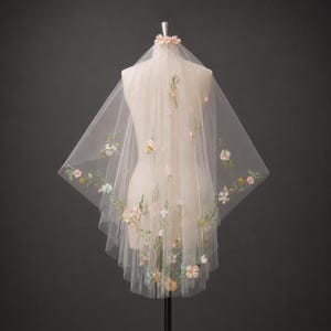 May include: A sheer, off-white tulle veil draped over a mannequin, featuring delicate, multi-colored floral embroidery. The embroidery includes small pink, blue, and yellow flowers with green stems and leaves scattered across the fabric.
