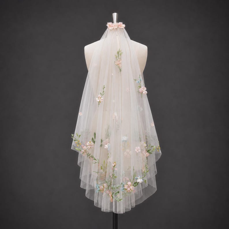 May include: A sheer, off-white tulle veil featuring delicate, multi-colored floral embroidery and small, scattered fabric flowers. The veil is gathered at the top with a small floral accent and drapes downward in a layered, semi-transparent design.