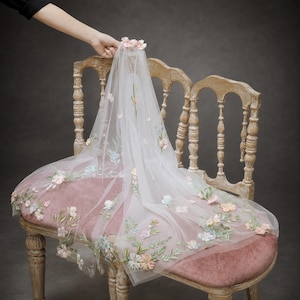 May include: A sheer white veil featuring delicate floral embroidery in shades of pink, blue, and green. The fabric is adorned with small, three-dimensional fabric flowers scattered across the surface, with a cluster of flowers at the top crown.
