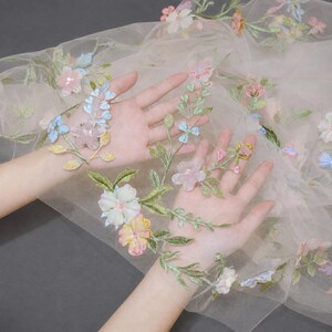 May include: A sheer, light beige tulle fabric featuring delicate, multi-colored floral embroidery. The design includes various flowers in shades of pink, blue, yellow, and white, accented with green stems and leaves scattered across the translucent material.