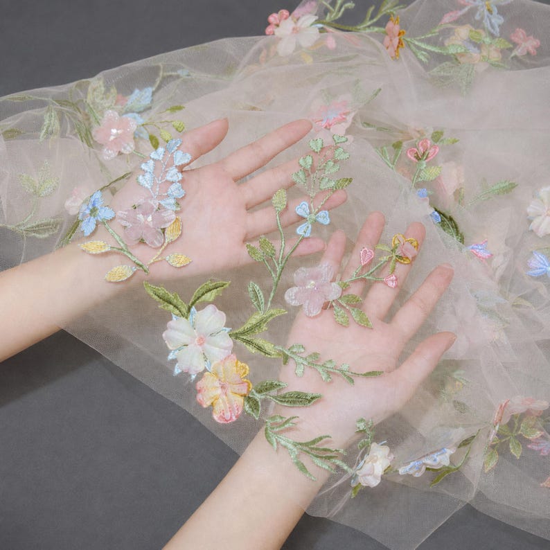May include: A sheer, light beige tulle fabric featuring delicate, multi-colored floral embroidery. The design includes various flowers in shades of pink, blue, yellow, and white, accented with green stems and leaves scattered across the translucent material.