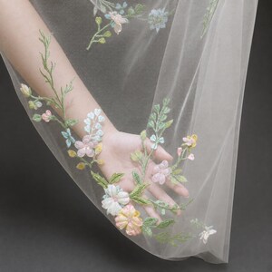 May include: A sheer white tulle fabric featuring delicate floral embroidery in soft pastel shades of pink, yellow, blue, and green. The intricate botanical pattern includes stems, leaves, and blossoms scattered across the translucent material.