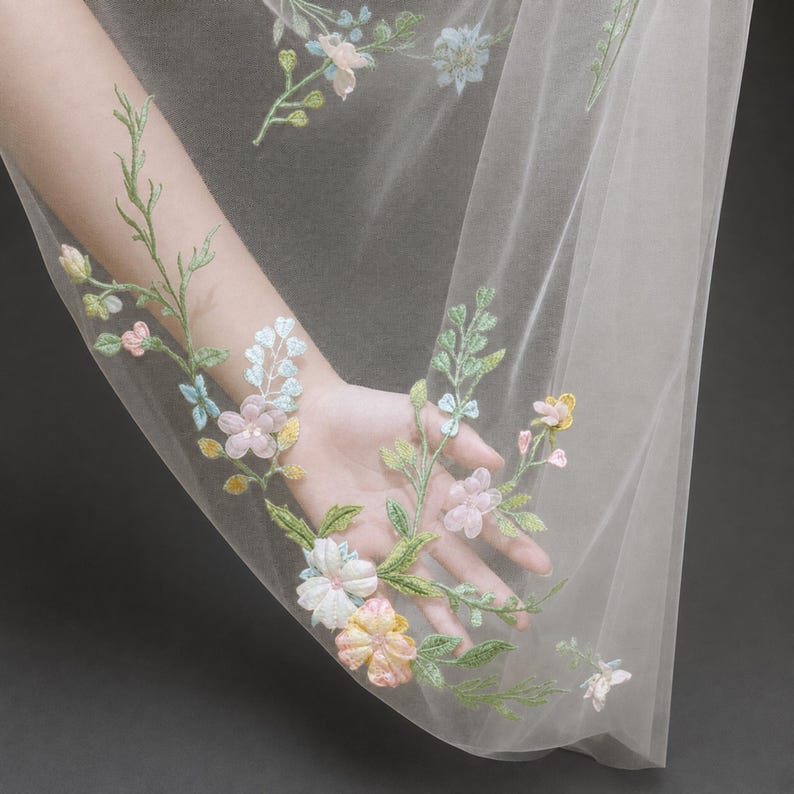May include: A sheer white tulle fabric featuring delicate floral embroidery in soft pastel shades of pink, yellow, blue, and green. The intricate botanical pattern includes stems, leaves, and blossoms scattered across the translucent material.