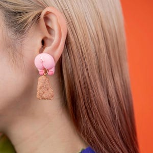 Puede incluir: Pendientes originales con forma de trasero rosa, con un conector dorado y un elemento rectangular marrón texturizado colgante. Los pendientes se llevan en una oreja, sobre un fondo naranja.