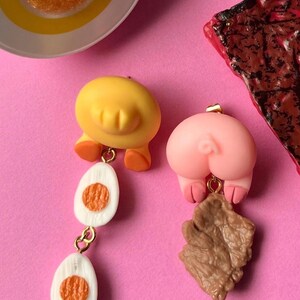 Puede incluir: Pendientes caprichosos. Un pendiente presenta una yema de huevo amarilla con pies naranjas y dos mitades de huevo colgantes, y un trasero de cerdo rosa con pies marrones sosteniendo un trozo de tocino. Sobre un fondo rosa.