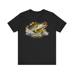 Pode incluir: Camiseta preta com uma ilustração detalhada de um peixe robalo saltando para fora da água. O design inclui a palavra "SNOOK" em letras em negrito, com "LINESIDER LEGEND" abaixo. O peixe é retratado com barbatanas amarelas e boca vermelha.