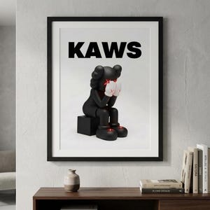 May include: Framed art print featuring a black KAWS figure seated on a black cube, with hands covering its face. The figure has red eyes and white gloves with an 'X' on each hand. The word 'KAWS' is printed in bold black letters at the top.
