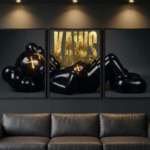 May include: A triptych wall art set featuring a glossy black character with glowing yellow X-shaped eyes. The central panel displays the word KAWS in large, textured gold lettering against a dark background, creating a cohesive three-piece display.