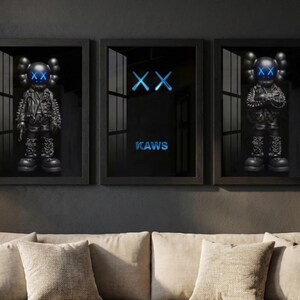 Peut inclure: Trois œuvres encadrées représentant des figurines KAWS et le logo. Les figurines noires ont des yeux bleus en forme de X et des tenues détaillées. Le cadre central affiche le logo KAWS en néon bleu sur fond noir. L'œuvre est au-dessus d'un canapé.