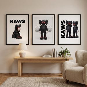 May include: Three framed art prints featuring KAWS figures. The prints showcase black figures with red 'XX' eyes and white gloves. One print has the word 'KAWS' in bold black letters. The prints are displayed above a wooden bench.