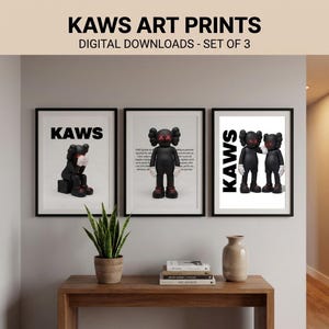 May include: Three framed KAWS art prints. Each print features a black KAWS figure with white gloves and red accents. The prints are displayed on a light-colored wall above a wooden table with a plant and decorative vase.