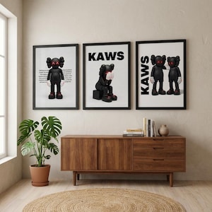 May include: Three framed art prints featuring KAWS characters. The prints have black frames and white backgrounds. The KAWS figures are black with red X eyes and hands. The word "KAWS" is printed in bold black letters on two of the prints.
