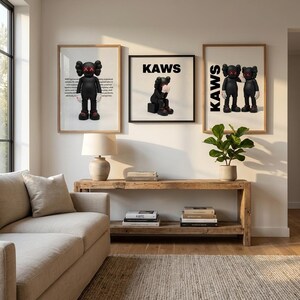 May include: Three framed KAWS art prints displayed on a wall. The prints feature black figures with X-shaped eyes and gloved hands. The word "KAWS" is printed in bold, black letters on two of the prints. The art is displayed above a wooden console table.