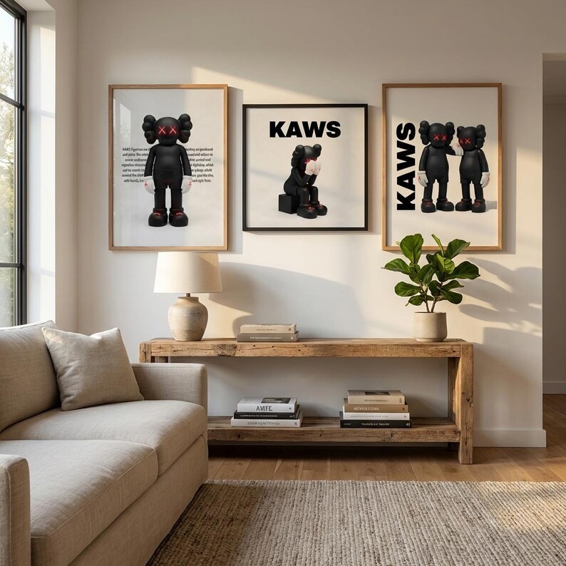 May include: Three framed KAWS art prints displayed on a wall. The prints feature black figures with X-shaped eyes and gloved hands. The word "KAWS" is printed in bold, black letters on two of the prints. The art is displayed above a wooden console table.
