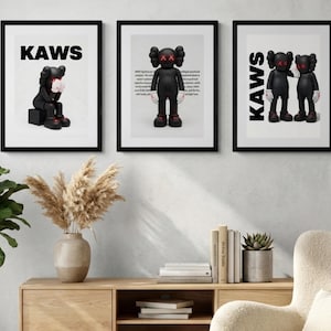 May include: Three framed art prints featuring black KAWS figures with red X eyes. The prints have the word "KAWS" in bold black letters at the top. The figures are in different poses, set against a white background.