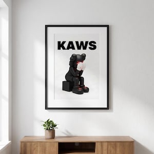 May include: Framed art print featuring a black figure with white gloves and red accents, seated on a black block. The word "KAWS" is printed in bold black letters above the figure. The artwork is displayed in a black frame.