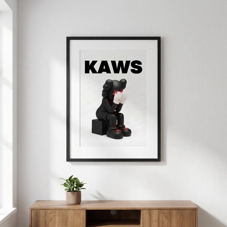 May include: Framed art print featuring a black figure with white gloves and red accents, seated on a black block. The word "KAWS" is printed in bold black letters above the figure. The artwork is displayed in a black frame.