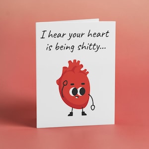 May include: A white greeting card featuring a cartoon illustration of a red anatomical heart with a face, arms, and legs. The text printed in black at the top of the card reads, I hear your heart is being shitty... The design is simple and graphic.