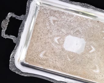 May include: Silver rectangular tray with ornate handles and a decorative floral pattern. The tray has a slightly raised edge and a rectangular center with a lighter color. The handles are curved and detailed.