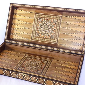 May include: A rectangular wooden backgammon board featuring intricate marquetry inlay. The interior displays light and dark wood triangular points and a central square panel with a geometric star pattern. The exterior edge has a decorative border.