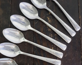 May include: Six silver spoons arranged diagonally on a dark wooden surface. The spoons have a classic design with elongated bowls and decorative handles. The spoons reflect the light, creating a bright contrast against the dark background. The spoons are likely for serving.