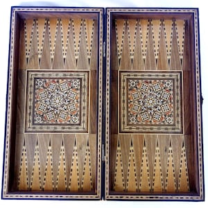 May include: Open backgammon board with a detailed inlaid design. The board is made of wood with a dark border and features a central square with a geometric star pattern. The playing surface has alternating light and dark triangles.