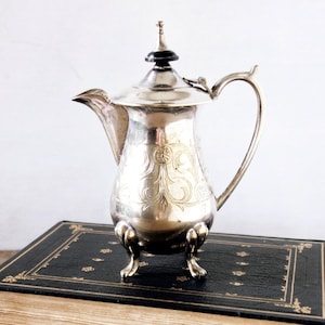 May include: A silver-colored, antique coffee pot with ornate floral engravings, a curved handle, and a black finial. The pot is displayed on a vintage book with gold detailing.